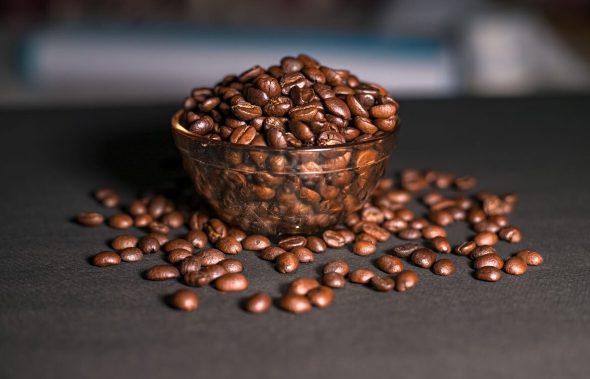 A clear bowl of whole coffee beans sits on a dark countertop.