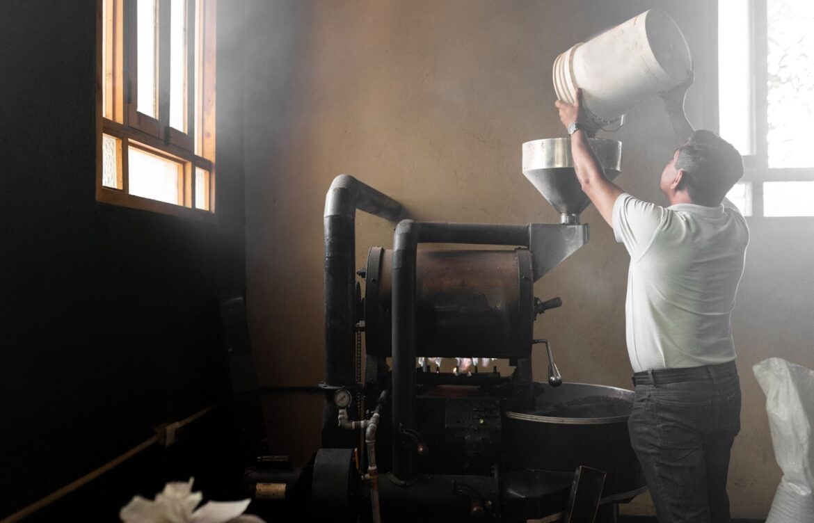 A man dumps a full bucket of coffee beans into a large roaster.